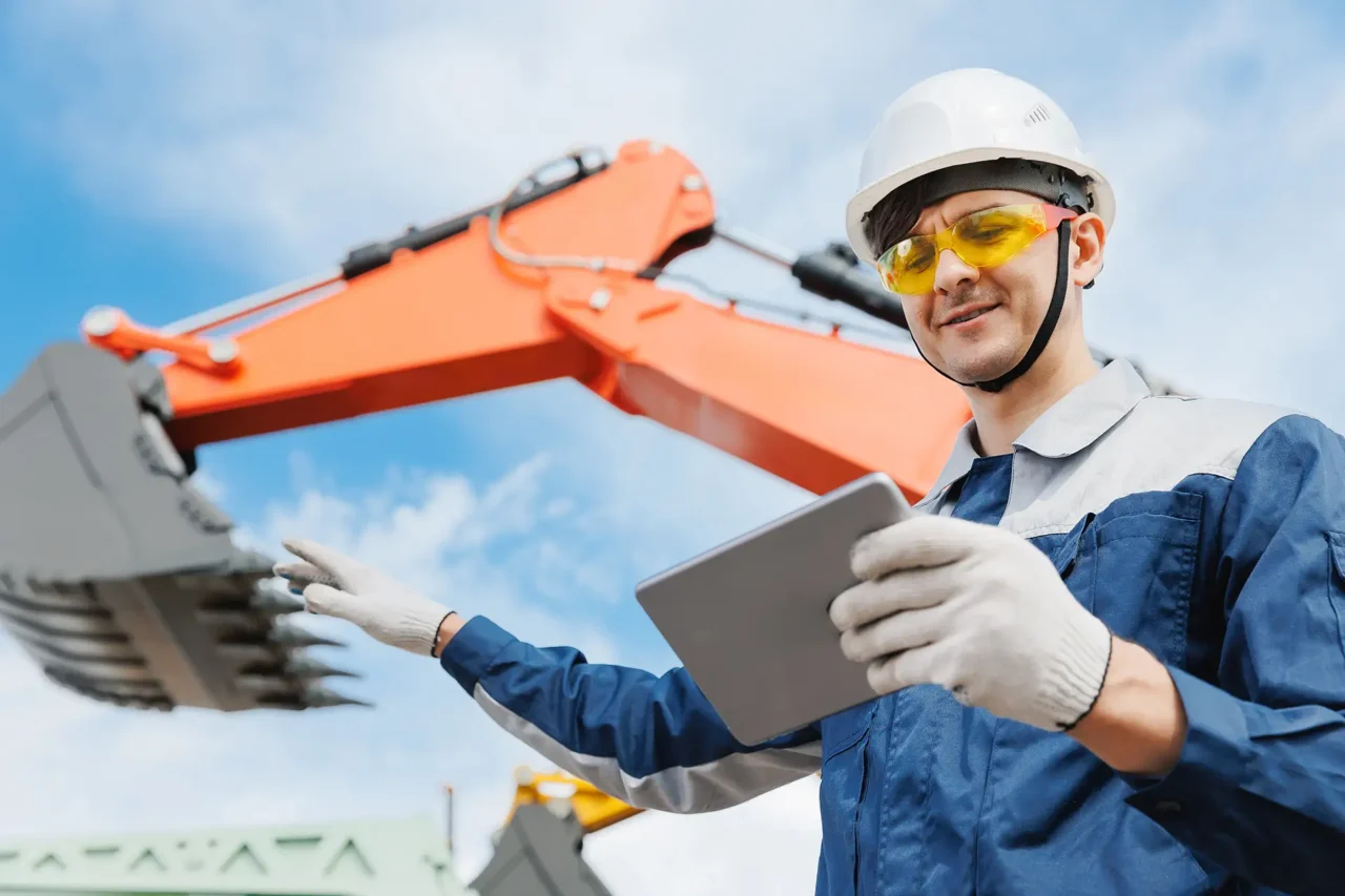Construction man looking at tablet with a orange digger behind him